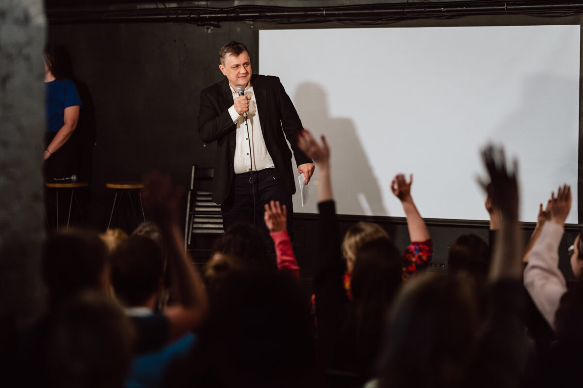 Nie mow do mnie ASAP Klub Komediowy 049 Ein Mann im Anzug spricht in ein Mikrofon auf einer Bühne vor einem sitzenden Publikum, von dem mehrere ihre Hände gehoben haben. Hinter ihm ist eine weiße Projektionsfläche zu sehen.
