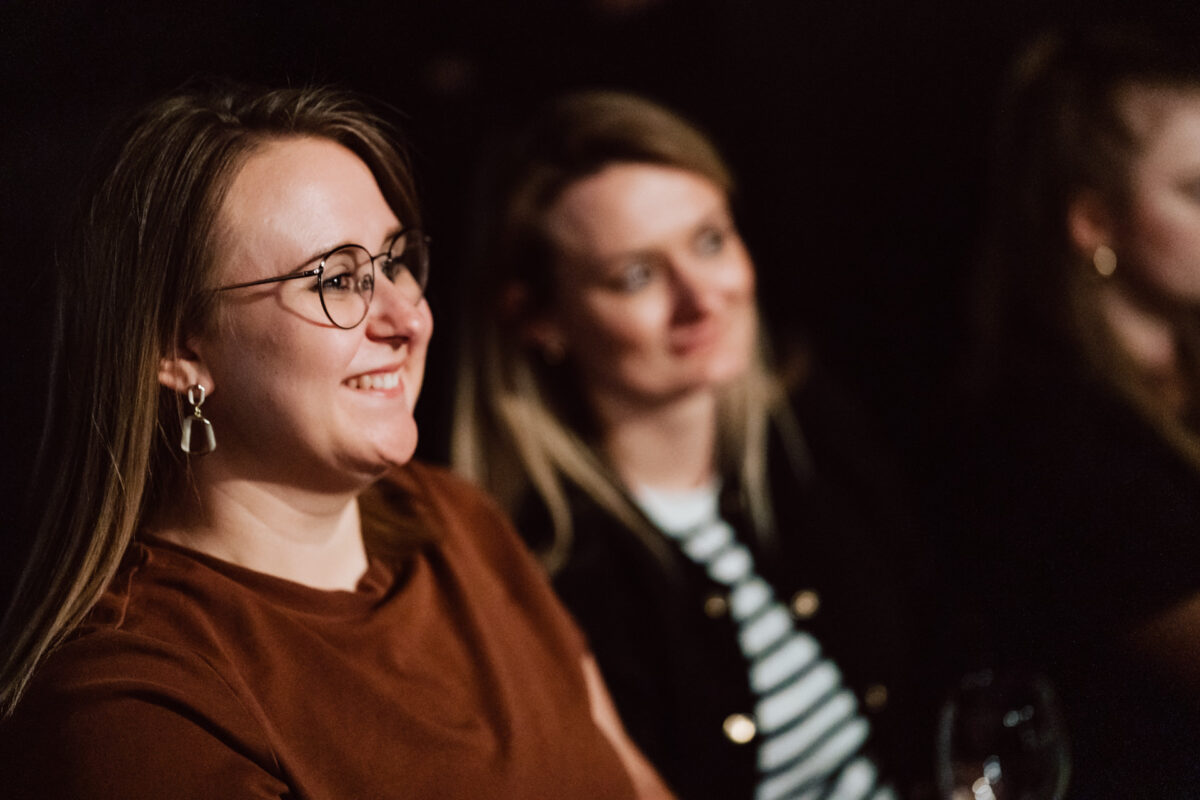Nie mow do mnie ASAP Klub Komediowy 050 Zwei Frauen sitzen Seite an Seite in einer schwach beleuchteten Umgebung. Die Frau im Vordergrund, die eine Brille und eine braune Bluse trägt, lächelt. Die Frau hinter ihr, in einem gestreiften Hemd und einer dunklen Jacke, ist leicht unscharf.