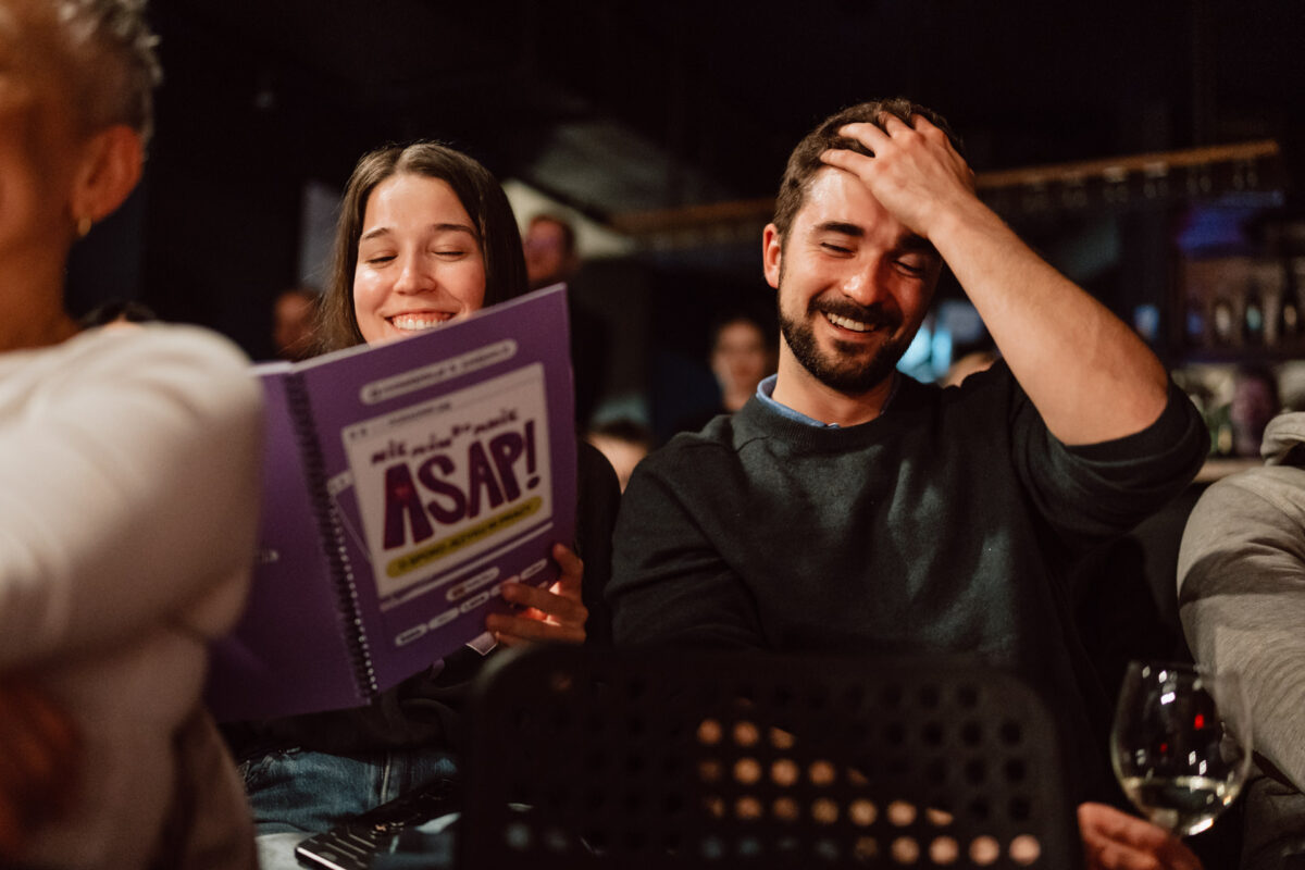Nie mow do mnie ASAP Klub Komediowy 051 Zwei Menschen sitzen dicht beieinander, lächeln und lachen. Eine von ihnen hält eine große lila Broschüre mit der Aufschrift "ASAP!" in der Hand, während die andere sich amüsiert an die Stirn fasst. Im Hintergrund sind weitere Personen und ein Glas Wein zu sehen.