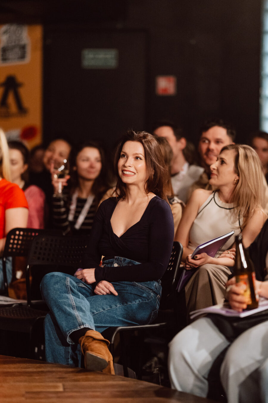 Nie mow do mnie ASAP Klub Komediowy 052 Eine Gruppe von Menschen sitzt im Publikum, lächelt und hört der Präsentation zu. Die Frau im Vordergrund trägt ein dunkles Oberteil und Jeans, hat die Beine gekreuzt und sieht engagiert und glücklich aus. Andere halten Getränke und Notizblöcke in der Hand.
