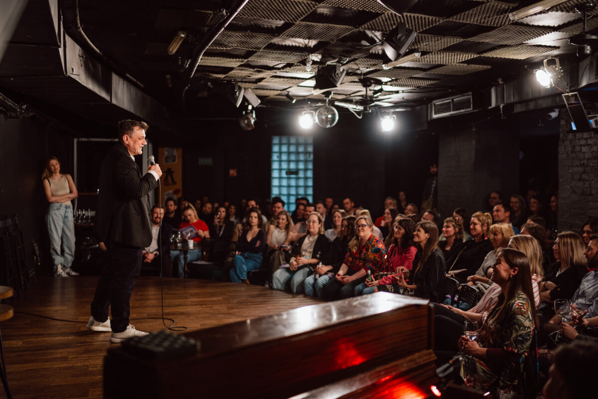 Nie mow do mnie ASAP Klub Komediowy 053 Der Comedian tritt auf der Bühne in einem schwach beleuchteten Comedy-Club auf. Er hält ein Mikrofon in der Hand, während ein großes Publikum dicht beieinander sitzt, zusieht und lacht. Der Raum hat dunkle Wände und Oberlichter.