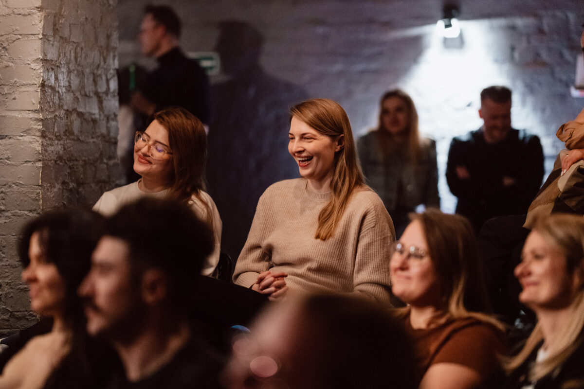 Nie mow do mnie ASAP Klub Komediowy 054 Eine Gruppe von Menschen sitzt dicht beieinander in einem schwach beleuchteten Raum, lächelt und lacht und beobachtet etwas aus dem Off. Die Stimmung scheint entspannt und fröhlich zu sein.
