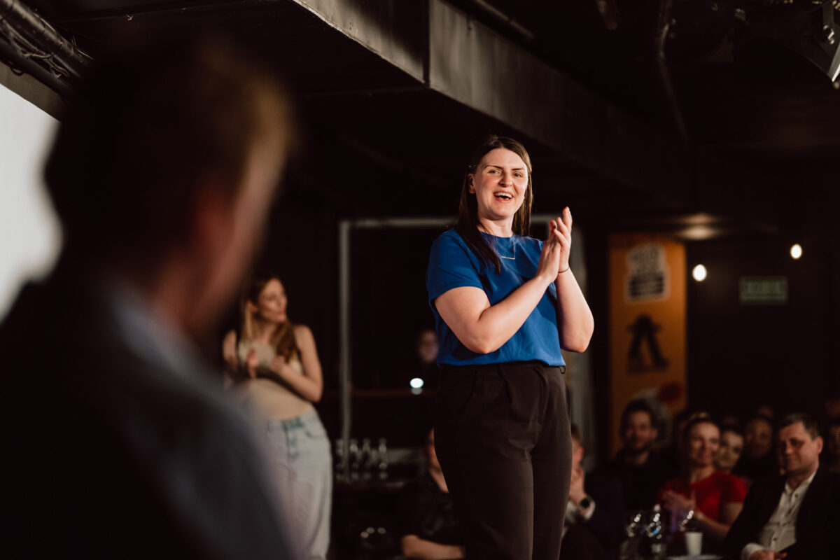 Nie mow do mnie ASAP Klub Komediowy 055 Eine Frau in einem blauen Hemd steht auf der Bühne, klatscht und lächelt das Publikum an. Die Leute im Hintergrund schauen zu und applaudieren in dem schwach beleuchteten Raum.