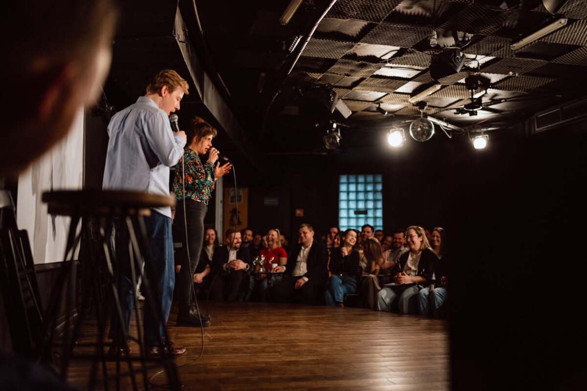 Nie mow do mnie ASAP Klub Komediowy 057 Zwei Personen stehen mit Mikrofonen auf der Bühne und treten vor einem aufmerksamen Publikum in einem schwach beleuchteten Comedy-Club oder Theater auf. Das Publikum sitzt dicht beieinander, schaut zu und lächelt. Auf der Bühne sind mehrere Barhocker zu sehen.