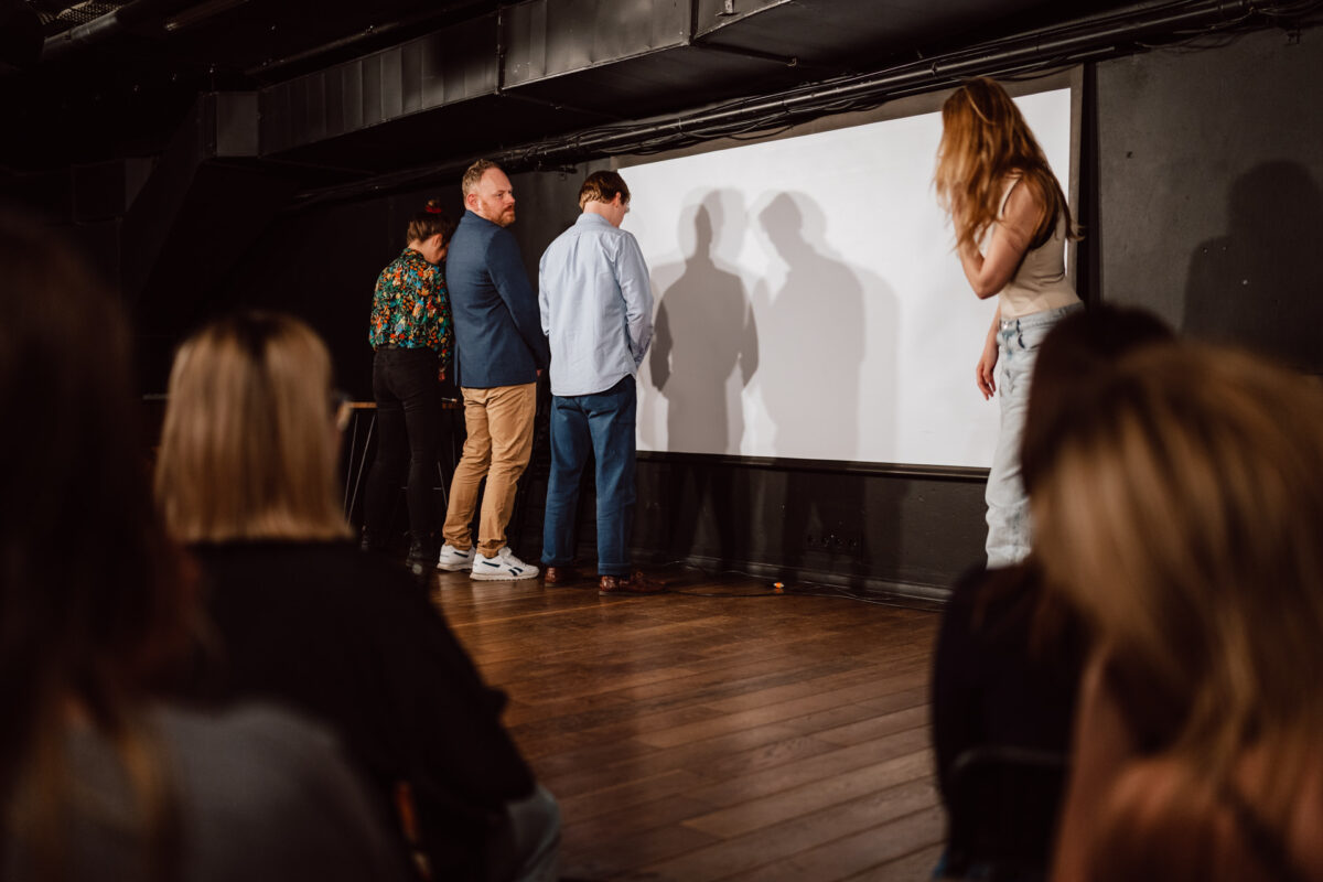 Nie mow do mnie ASAP Klub Komediowy 060 Vier Personen stehen vor einem weißen Bildschirm auf der Bühne, während eine Frau rechts daneben steht und zuschaut und das Publikum im Vordergrund zusieht. Es scheint sich um eine Aufführung oder einen Workshop in einem schwach beleuchteten Raum zu handeln.