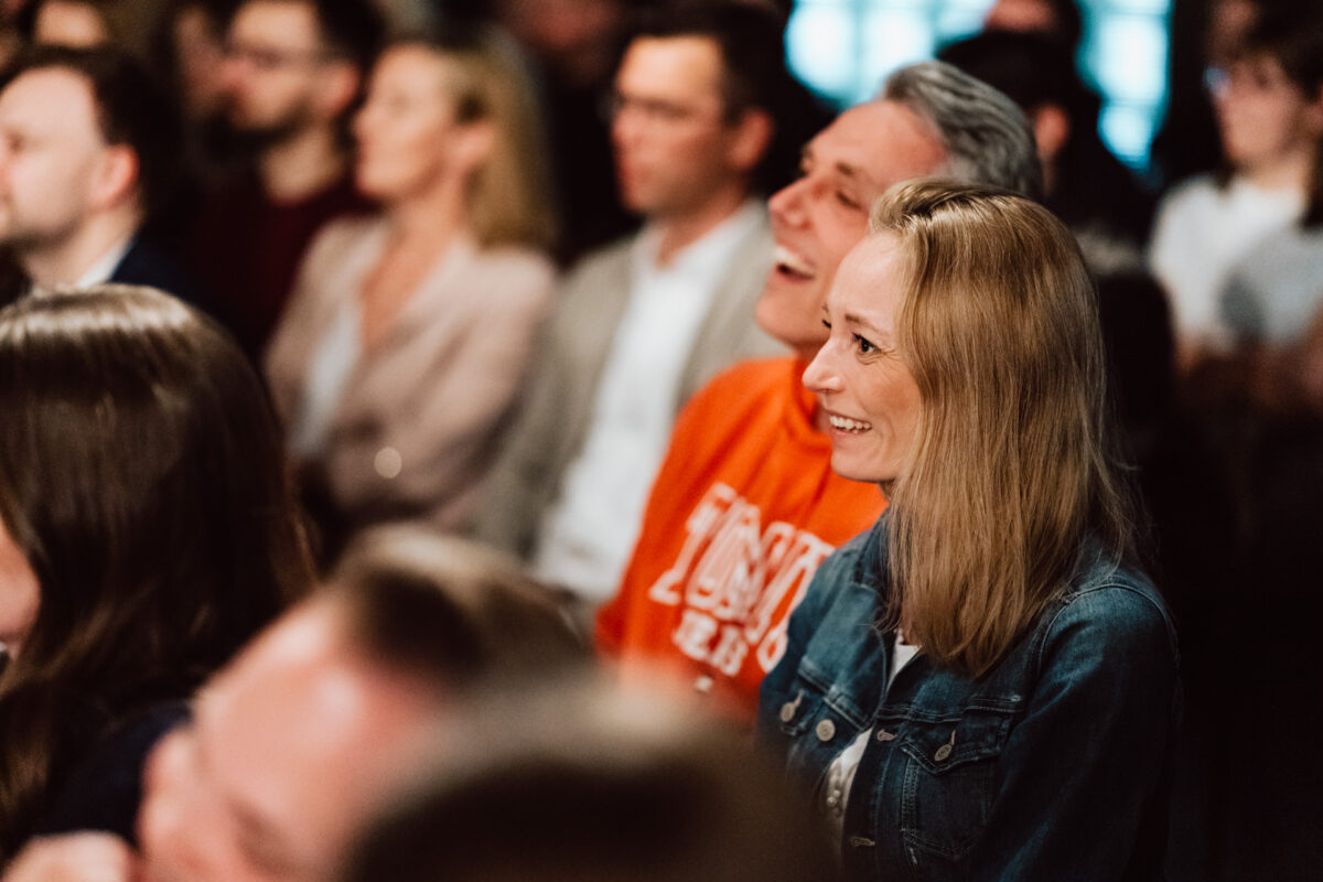 Nie mow do mnie ASAP Klub Komediowy 062 Eine Gruppe von Menschen sitzt bei einer Indoor-Party dicht beieinander, lächelt und sieht beschäftigt aus. Der Fokus liegt auf einer Frau in einer Jeansjacke im Vordergrund, während andere Personen im Hintergrund verschwommen sind.