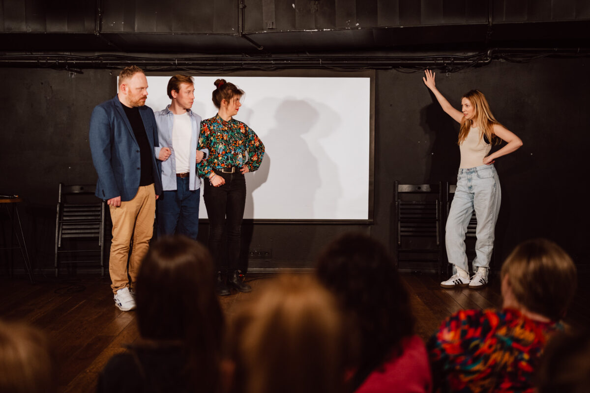 Nie mow do mnie ASAP Klub Komediowy 063 Vier Personen stehen vor einem weißen Bildschirm auf der Bühne und blicken auf eine Frau in Jeans und T-Shirt, die mit erhobener Hand rechts steht. Das Publikum schaut vom Vordergrund aus zu.