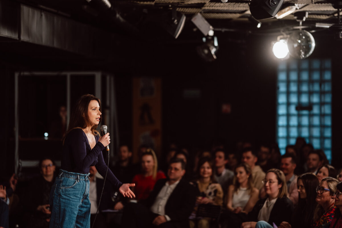 Nie mow do mnie ASAP Klub Komediowy 066 Eine Frau steht auf der Bühne und hält ein Mikrofon in der Hand. Sie tritt vor einem aufmerksamen Publikum auf, das in einem schwach beleuchteten Comedy-Club oder Veranstaltungsraum sitzt.