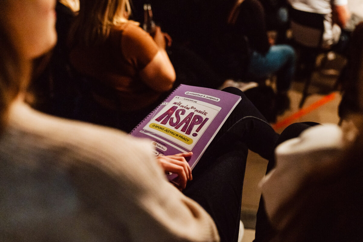 Nie mow do mnie ASAP Klub Komediowy 070 Eine Person sitzt mit einem Spiralnotizbuch auf dem Schoß. Auf dem lilafarbenen Einband des Notizbuchs steht in fetten Buchstaben "ASAP!", während im Hintergrund Menschen dicht beieinander sitzen.