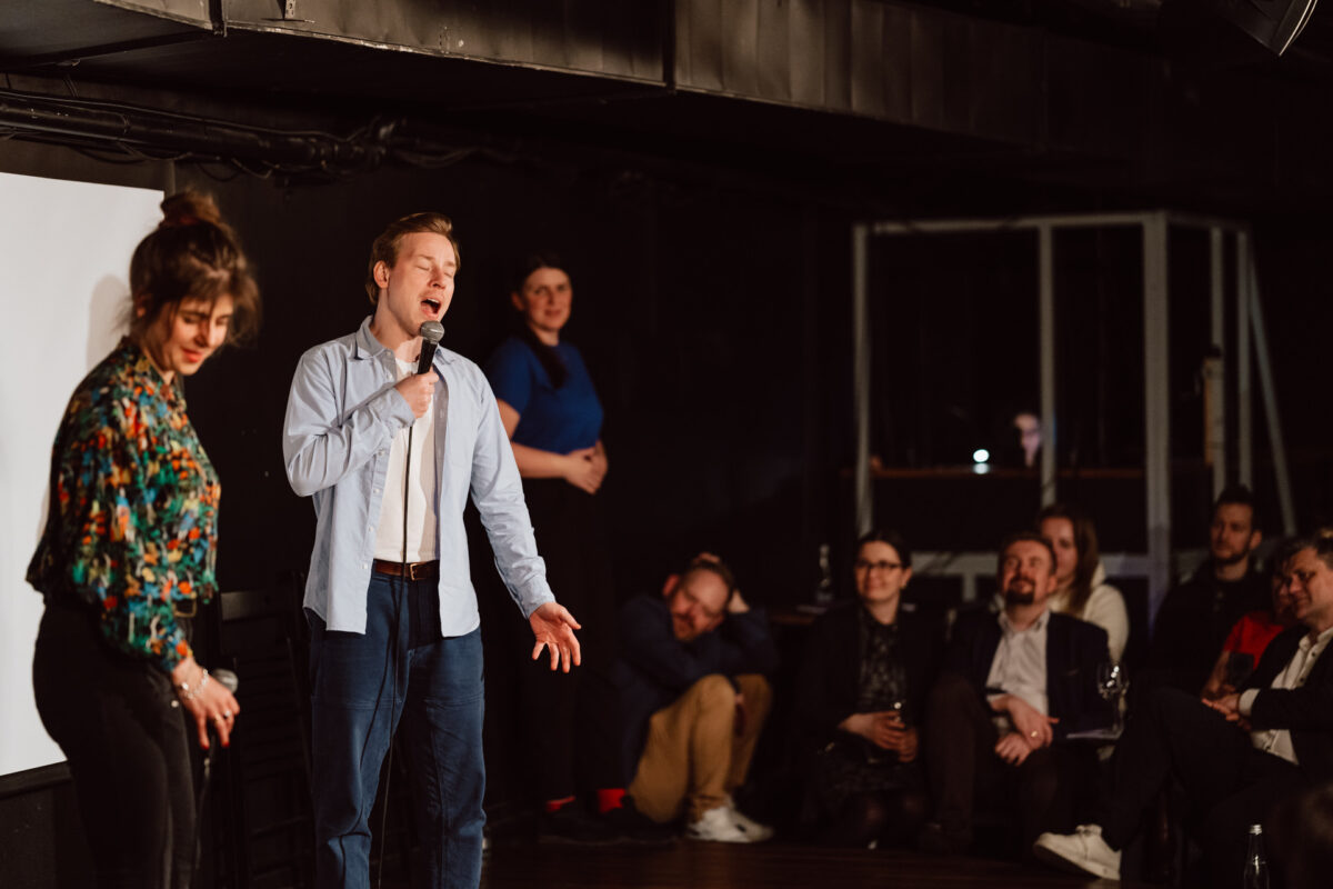 Nie mow do mnie ASAP Klub Komediowy 072 Ein Mann singt auf der Bühne leidenschaftlich in ein Mikrofon, während eine Frau neben ihm nach unten schaut. Mehrere Menschen sitzen und stehen im schwach beleuchteten Publikum und schauen aufmerksam zu.