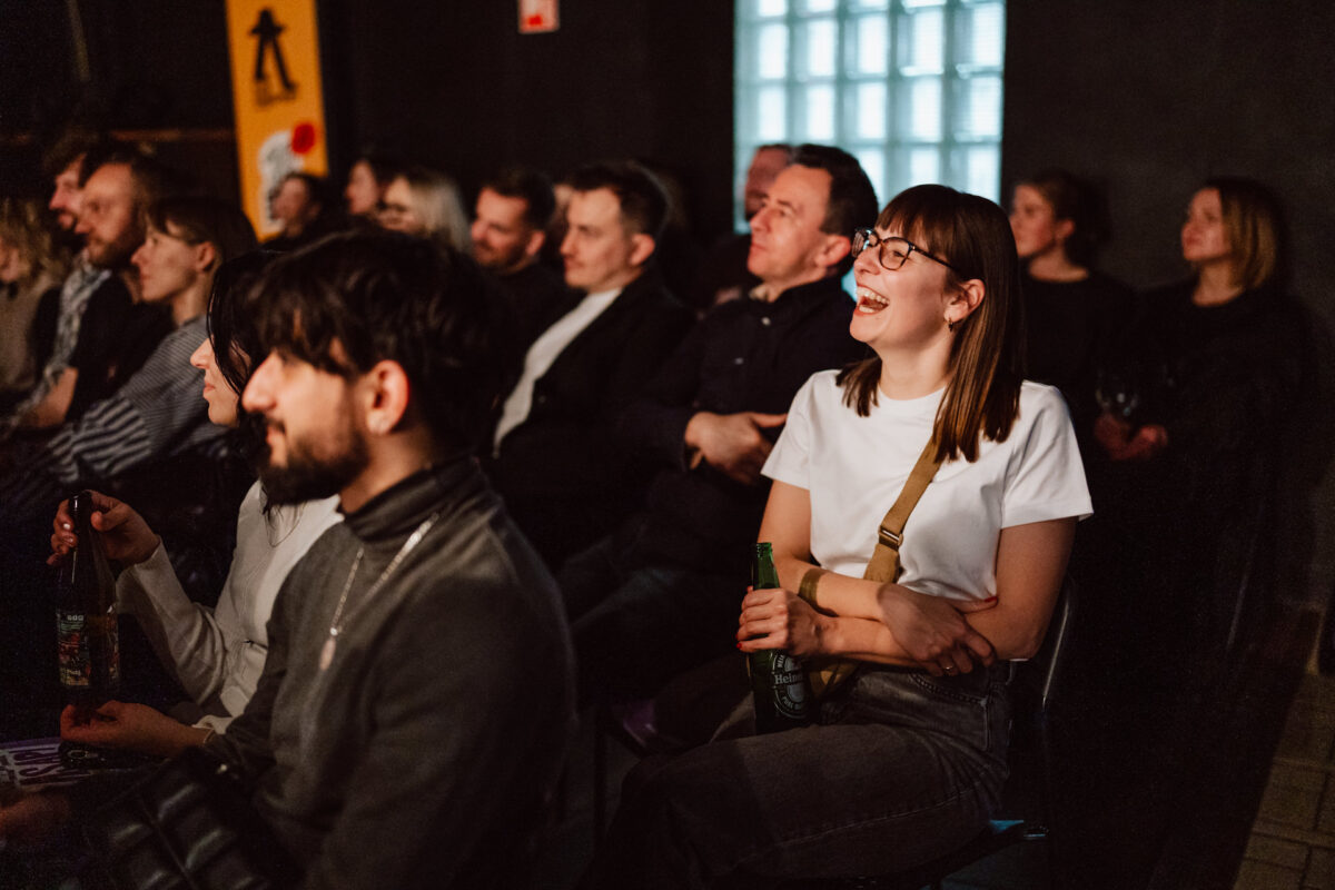 Nie mow do mnie ASAP Klub Komediowy 075 Das Publikum sitzt dicht gedrängt auf einer Indoor-Party. Die meisten Leute schauen aufmerksam zu, während eine Frau in einem weißen Hemd lächelt und lacht, während sie ein Getränk in der Hand hält. Die Stimmung scheint entspannt und fröhlich zu sein.