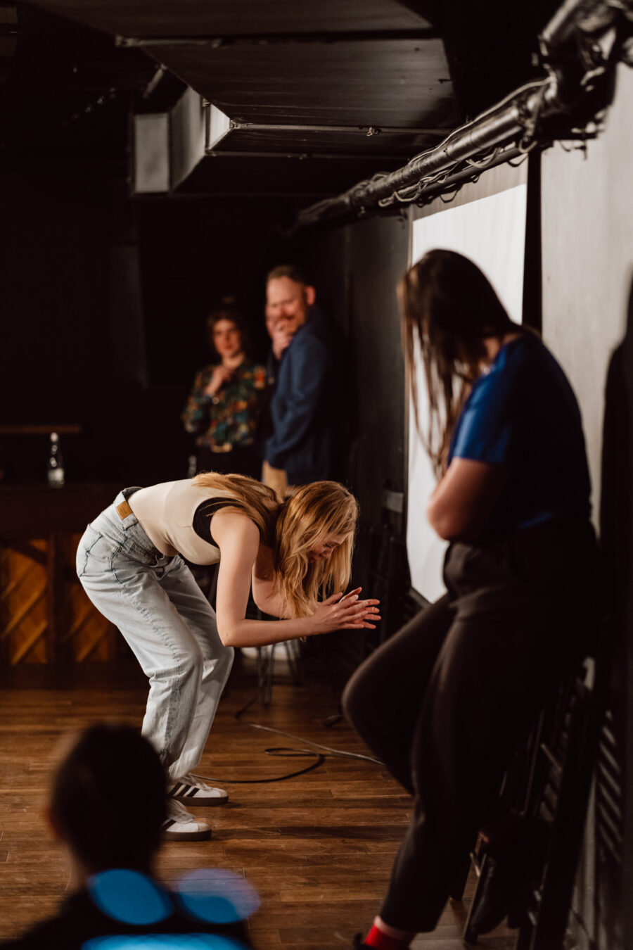 Nie mow do mnie ASAP Klub Komediowy 077 Eine Frau in einem weißen T-Shirt und Jeans lehnt sich mit gefalteten Händen vor, während andere sie auf einer schwach beleuchteten Bühne beobachten. Zwei Personen stehen im Hintergrund und eine weitere Person in einem blauen Outfit steht im Vordergrund.