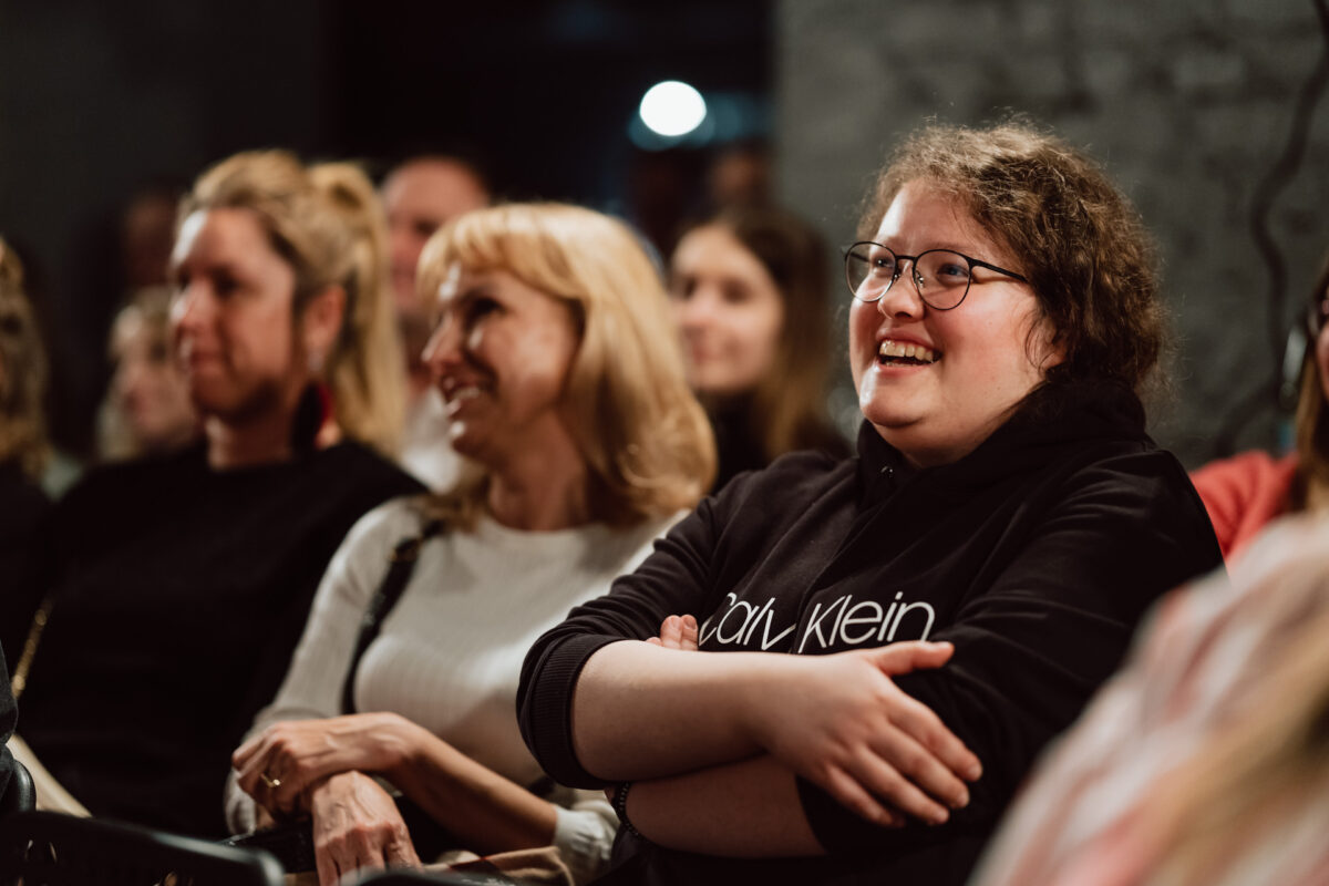 Nie mow do mnie ASAP Klub Komediowy 081 Eine Gruppe von Menschen, meist Frauen, sitzt dicht beieinander im Publikum. Sie lächeln und scheinen engagiert zu sein. Eine Frau im Vordergrund trägt eine Brille und einen dunklen Kapuzenpulli, hat die Arme verschränkt und genießt die Veranstaltung.