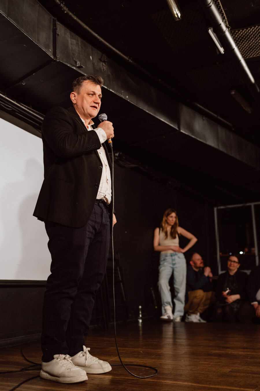 Nie mow do mnie ASAP Klub Komediowy 082 Ein Mann in einer schwarzen Jacke und weißen Turnschuhen hält auf der Bühne ein Mikrofon und spricht zum Publikum. Im Hintergrund sehen eine Frau und drei sitzende Personen zu und lächeln. Es scheint sich um eine Komödie oder eine Aufführung zu handeln.