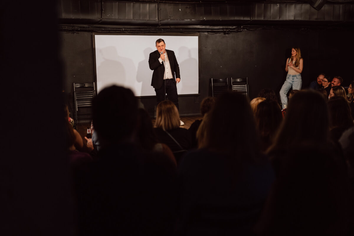 Nie mow do mnie ASAP Klub Komediowy 083 Ein Mann im Anzug tritt auf der Bühne mit einem Mikrofon als Stand-up-Comedian auf, während das Publikum zusieht und eine Frau an der Seite steht, lächelt und klatscht. Der Hintergrund ist dunkel und hinter ihm befindet sich eine weiße Leinwand.