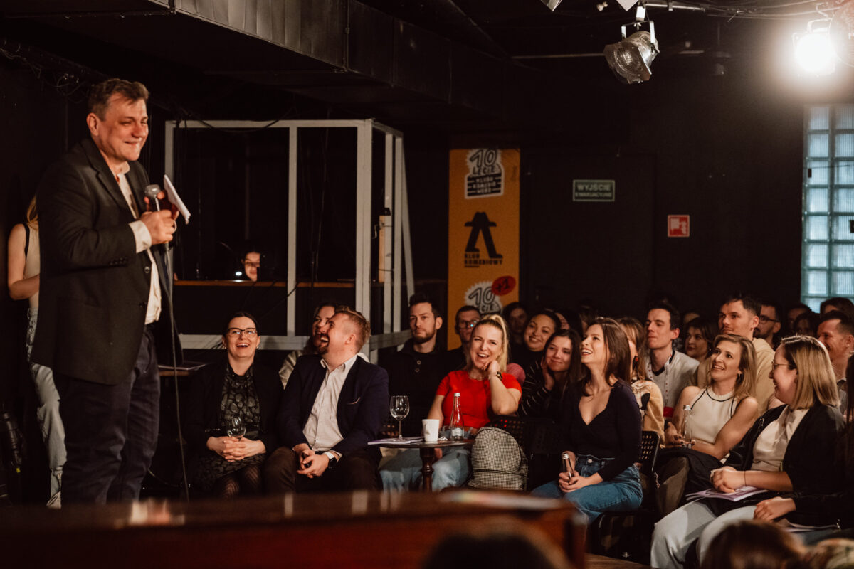 Nie mow do mnie ASAP Klub Komediowy 085 Ein Mann mit einem Mikrofon tritt auf der Bühne eines schummrig beleuchteten Comedy-Clubs vor einem sitzenden Publikum auf. Das Publikum lächelt und lacht und auf den Tischen stehen Getränke. Die Atmosphäre scheint lebhaft und einnehmend zu sein.