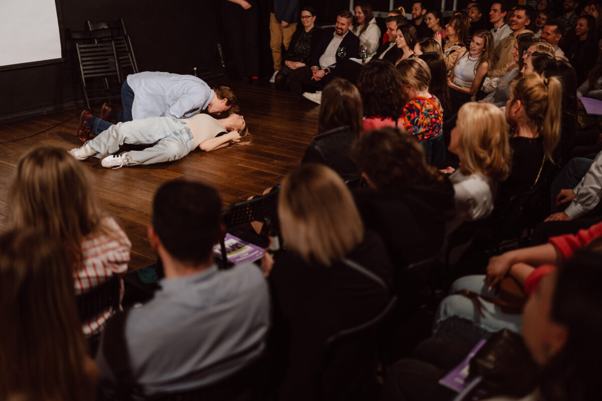 Nie mow do mnie ASAP Klub Komediowy 091 Der Darsteller liegt auf dem Holzboden der Bühne und eine weitere Person lehnt sich über ihn, beide in legerer Kleidung, während ein großes Publikum in der Theaterkulisse zusieht und lacht.