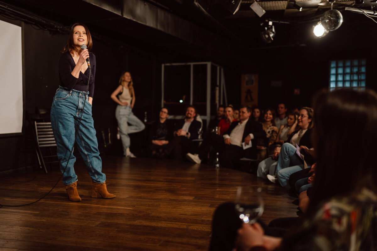 Nie mow do mnie ASAP Klub Komediowy 096 Eine Frau, die ein Mikrofon in der Hand hält, macht Stand-up-Comedy auf der Bühne vor einem sitzenden Publikum in einem schwach beleuchteten Comedy-Club. Das Publikum sieht engagiert und amüsiert aus.