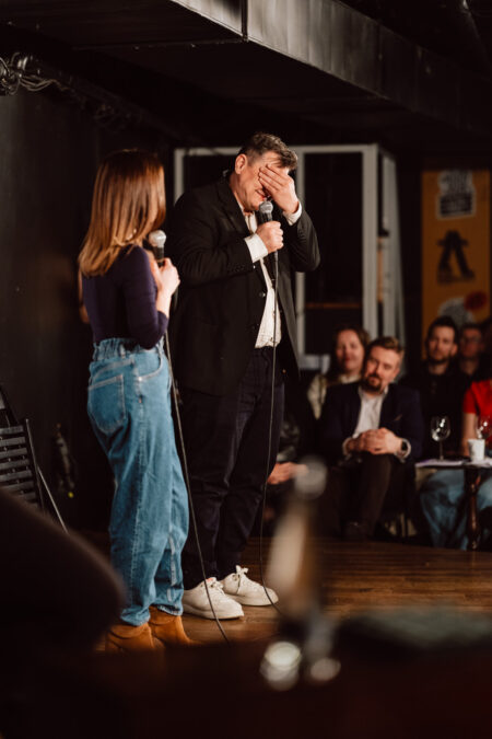 Nie mow do mnie ASAP Klub Komediowy 099 Ein Mann auf der Bühne bedeckt sein Gesicht mit einer Hand, während er ein Mikrofon hält, und steht neben einer Frau mit einem Mikrofon. Mehrere Personen sitzen in einem schwach beleuchteten Raum und beobachten sie vom Publikum aus.