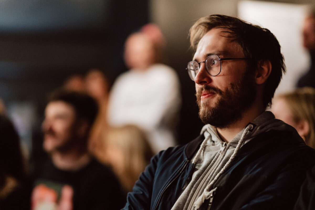 Nie mow do mnie ASAP Klub Komediowy 102 Ein Mann mit Brille und Bart, der ein Kapuzensweatshirt und eine Jacke trägt, sitzt inmitten eines verschwommenen Publikums und starrt konzentriert nach vorne. Der Hintergrund ist unscharf.