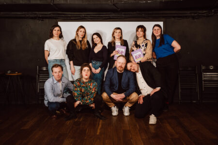 A group of eleven adults pose in a room on a wooden floor with a dark background. Three men and one woman kneel in front, and seven women stand behind them. Two women in the back row hold purple pamphlets and all look into the camera.