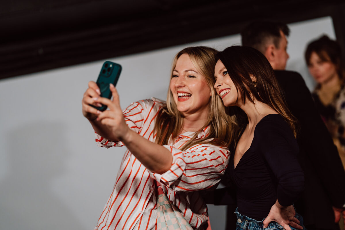 Nie mow do mnie ASAP Klub Komediowy 106 Zwei Frauen lächeln und posieren für ein gemeinsames Selfie, eine hält ein Smartphone. Andere Personen sind unscharf im Hintergrund zu erkennen, was auf ein geselliges oder zwangloses Beisammensein schließen lässt.