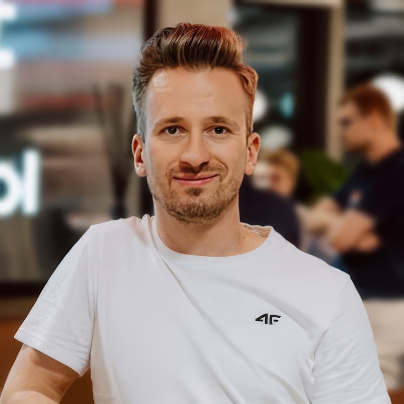 Marcin Krokowski fotograf eventowy links Ein Mann mit kurzem, gestyltem Haar und gestutztem Bart trägt ein weißes T-Shirt und lächelt in die Kamera. Der Hintergrund ist unscharf und zeigt andere Menschen und eine moderne Einrichtung.