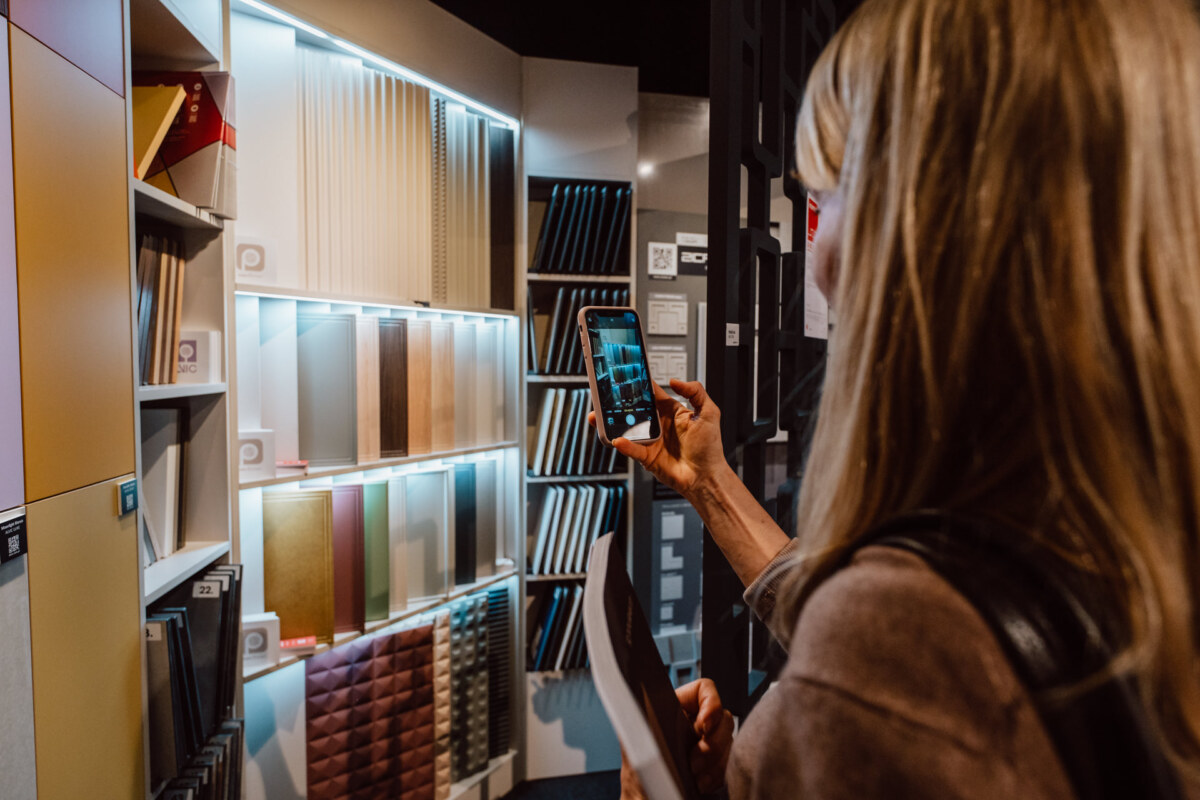 reportaz targi architektura architectatwork 2025 1 Eine Person mit langen blonden Haaren fotografiert farbenfrohe Materialmuster, die in einem Geschäft in den Regalen ausgestellt sind, und hält ein Telefon und eine Broschüre oder ein Muster in der Hand.