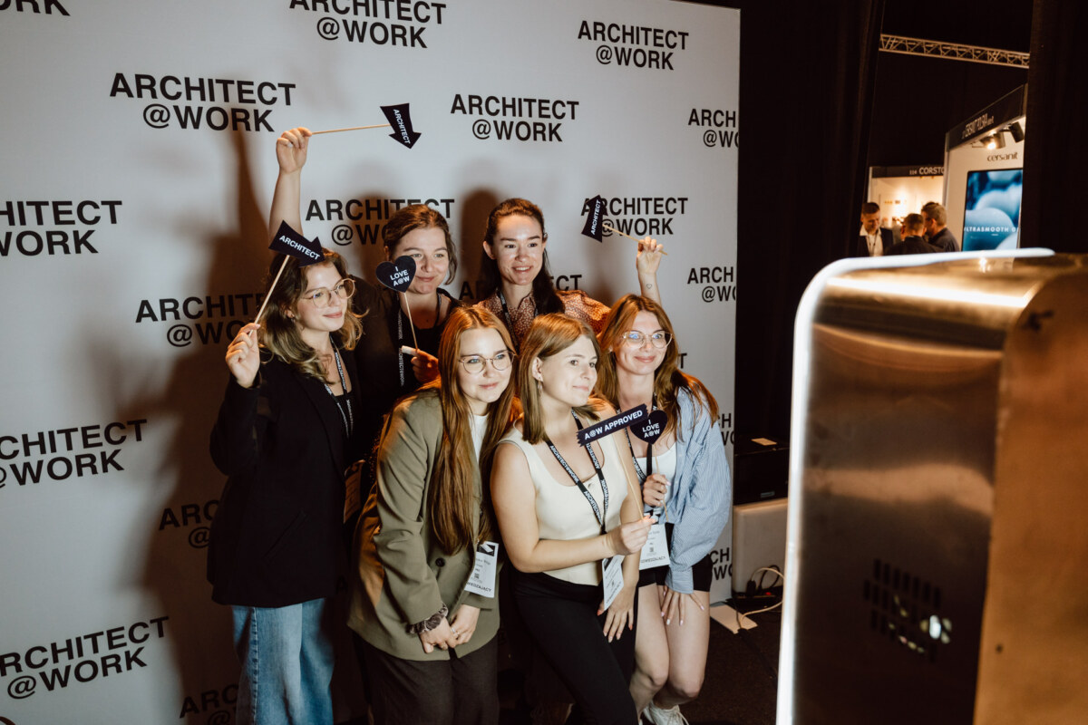 reportaz targi architektura architectatwork 2025 10 Sechs Frauen posieren und lächeln vor dem Hintergrund von 'ARCHITECT @ WORK', einige halten kleine Schilder, während sie ein Gruppenfoto auf der Veranstaltung machen.