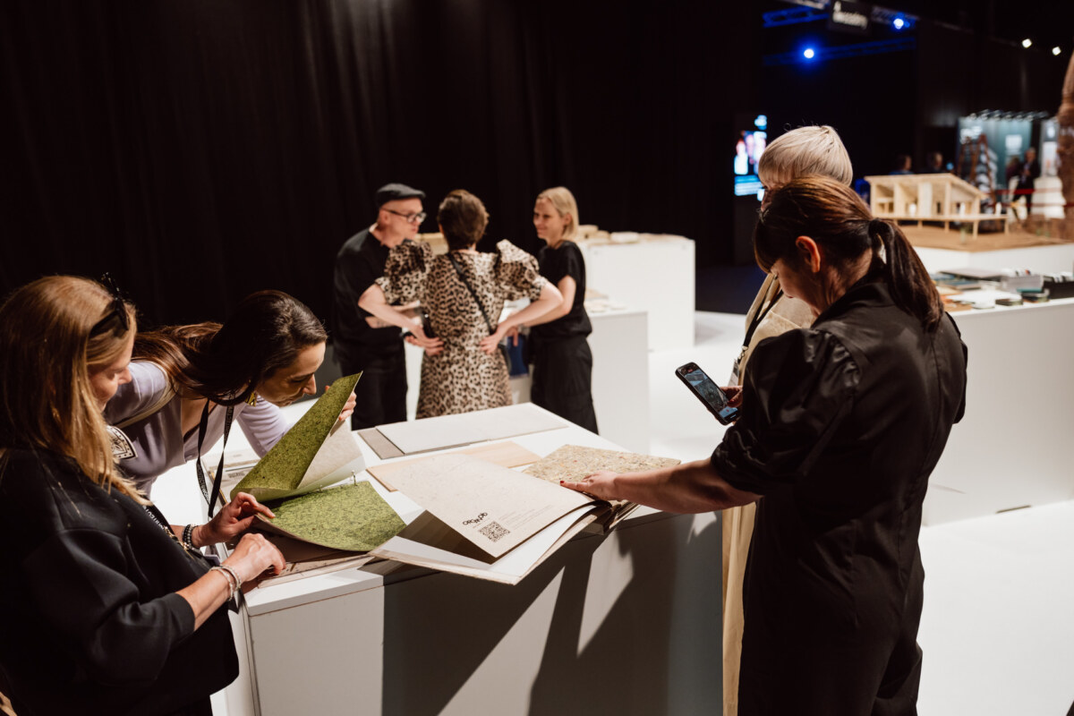 reportaz targi architektura architectatwork 2025 100 In einem schwach beleuchteten Ausstellungsraum stehen mehrere Personen um Ausstellungstische herum und untersuchen aufgeschlagene Bücher oder strukturierte Materialien. Einige lesen aufmerksam, während eine Person ein Smartphone benutzt. Der Hintergrund ist dunkel und modern.