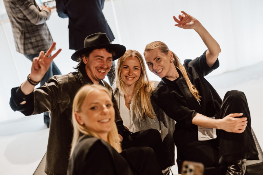 reportaz targi architektura architectatwork 2025 101 Vier lächelnde Menschen, drei Frauen und ein Mann, sitzen dicht beieinander und posieren für ein lustiges Gruppen-Selfie. Sie sind drinnen, stilvoll gekleidet, machen Friedenszeichen und feierliche Gesten.