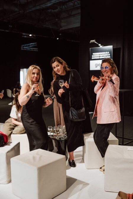 Drei Frauen stehen zusammen in einem Raum, halten Gläser und lächeln in die Kamera. Sie sind stilvoll gekleidet und scheinen an einer gesellschaftlichen Veranstaltung oder einem Networking-Event teilzunehmen, umgeben von weißen Würfeln und Menschen im Hintergrund. 