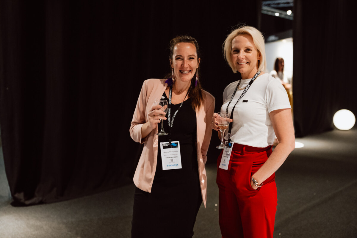 reportaz targi architektura architectatwork 2025 103 Zwei lächelnde Frauen, die bei der Veranstaltung Getränke in der Hand halten, beide mit Namensschildern; die eine in einem schwarzen Kleid mit rosa Jacke, die andere in einem weißen Hemd und einer roten Hose, stehen in einem schwach beleuchteten Raum mit schwarzen Vorhängen.