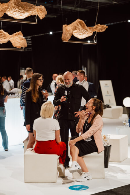 Eine Gruppe von Menschen in Geschäftskleidung trifft sich und unterhält sich bei einer modernen Indoor-Veranstaltung. Sie stehen und sitzen auf weißen Möbeln unter kunstvollen Hängedekorationen, während im Hintergrund andere Personen und Displays zu sehen sind.
