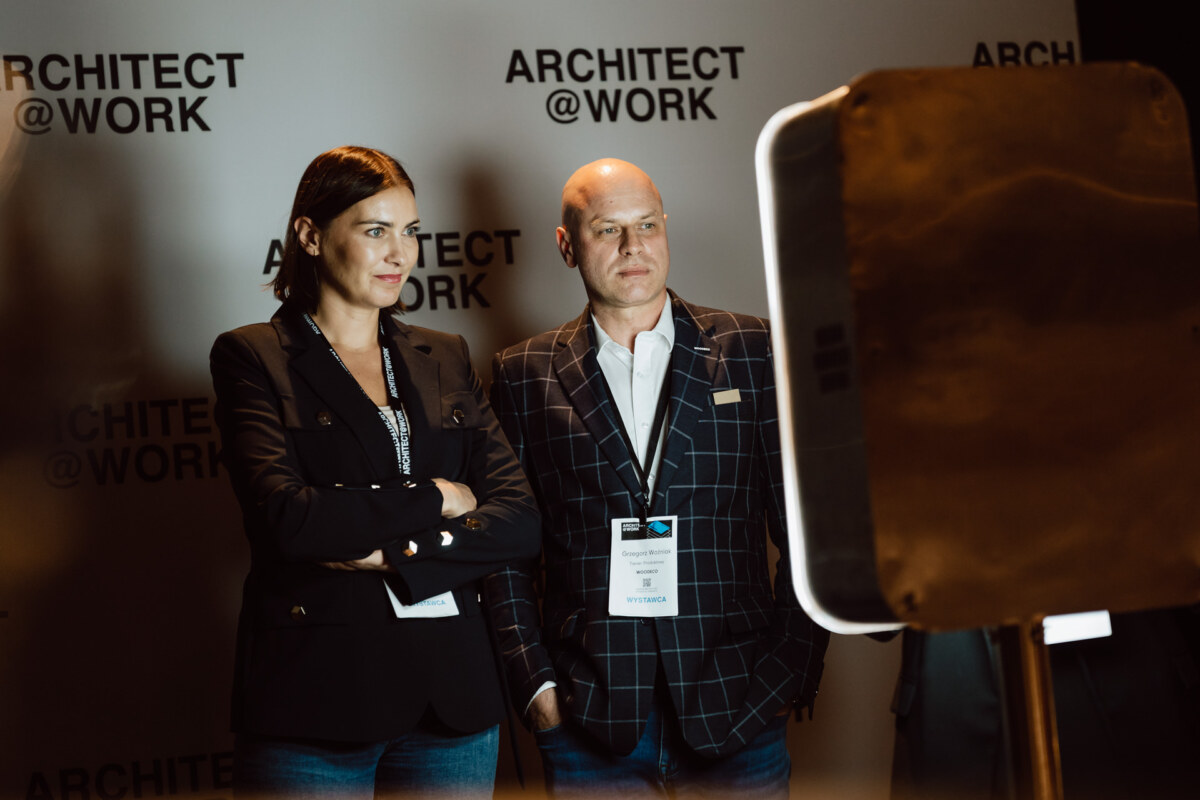 reportaz targi architektura architectatwork 2025 106 Zwei Personen mit Konferenzabzeichen stehen vor einem Hintergrund mit der Aufschrift 'ARCHITECT @ WORK' und schauen auf einen Bildschirm oder ein Gerät im Vordergrund. Die Atmosphäre wirkt professionell und erinnert an eine Veranstaltung.