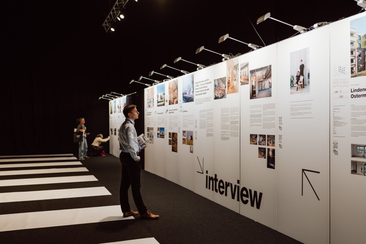 reportaz targi architektura architectatwork 2025 11 Eine Person steht lesend vor einer großen weißen Ausstellungswand mit Fotos und Text in hellem Licht in einem modernen Galerieraum. Auch andere Besucher beobachten die Auslagen im Hintergrund.