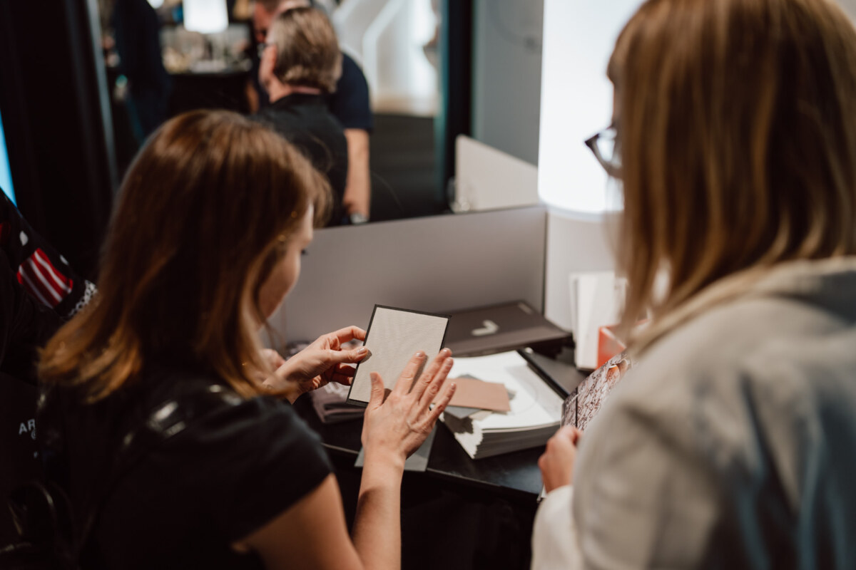 reportaz targi architektura architectatwork 2025 12 Zwei Frauen betrachten Materialproben an einem Tisch, eine von ihnen hält ein quadratisches Stück. Sie scheinen mit anderen Personen im Hintergrund über Gestaltungsmöglichkeiten in einem modernen Interieur zu diskutieren.