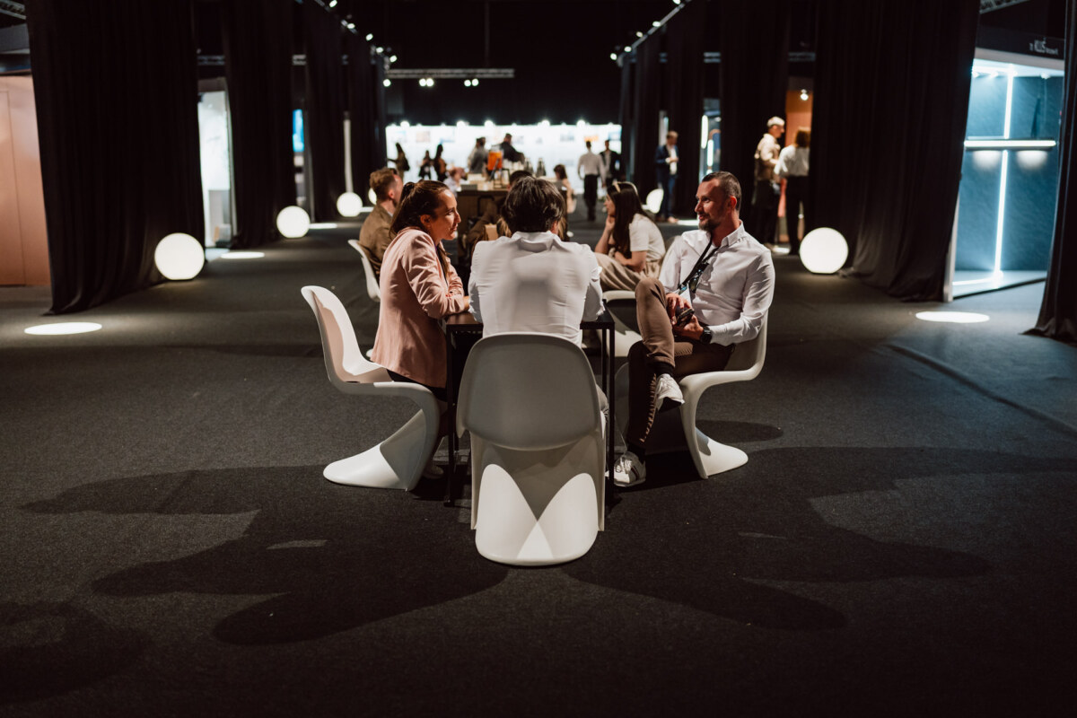 reportaz targi architektura architectatwork 2025 14 Mehrere Personen in Geschäftskleidung sitzen um moderne weiße Stühle an einem Tisch in einem schwach beleuchteten Konferenz- oder Veranstaltungsraum mit schwarzen Vorhängen und leuchtenden runden Lichtern im Hintergrund.