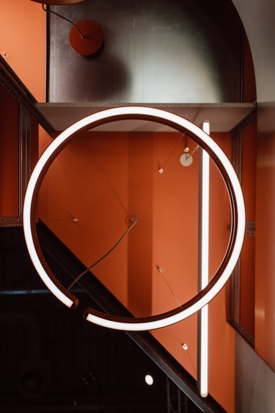 Eine große runde Leuchte hängt von der Decke vor einem orangefarbenen und metallenen industriellen Hintergrund und schafft ein modernes abstraktes Design mit geometrischen Formen und klaren Linien.