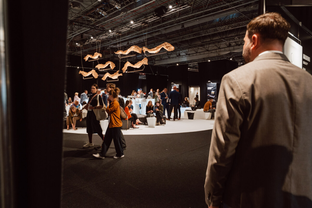 reportaz targi architektura architectatwork 2025 16 Ein Mann im Anzug steht im Vordergrund und beobachtet die Menschen, die sich in einem modernen, gut beleuchteten Veranstaltungsraum mit kunstvollen Lichtern über dem Kopf und Gruppen von Teilnehmern, die sitzen und sich mischen, versammeln.