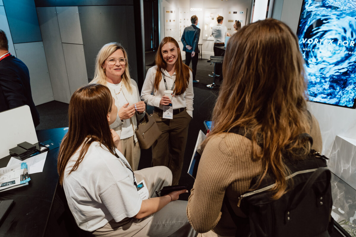 reportaz targi architektura architectatwork 2025 17 Vier Frauen stehen und sitzen zusammen, lächeln und unterhalten sich auf einer scheinbar professionellen Veranstaltung oder Konferenz. Sie tragen Abzeichen und legere Geschäftskleidung in einem modernen Interieur.