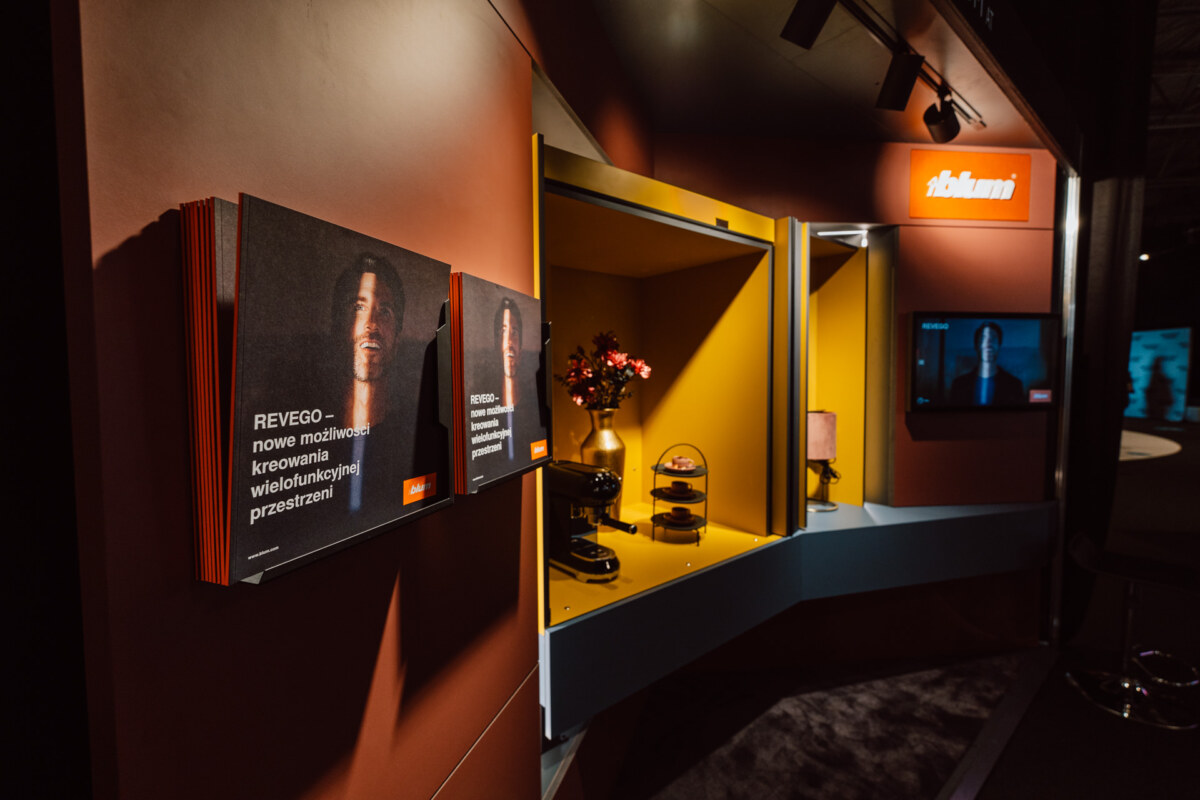 reportaz targi architektura architectatwork 2025 18 Ein moderner Ausstellungsstand mit orangefarbenen und gelben Wänden zeigt aufgeschlagene Bücher, eine Blumenvase, dekorative Gegenstände und einen Bildschirm, auf dem eine Person abgebildet ist. Der Stand hat ein 'Rikami'-Logo in der oberen rechten Ecke.