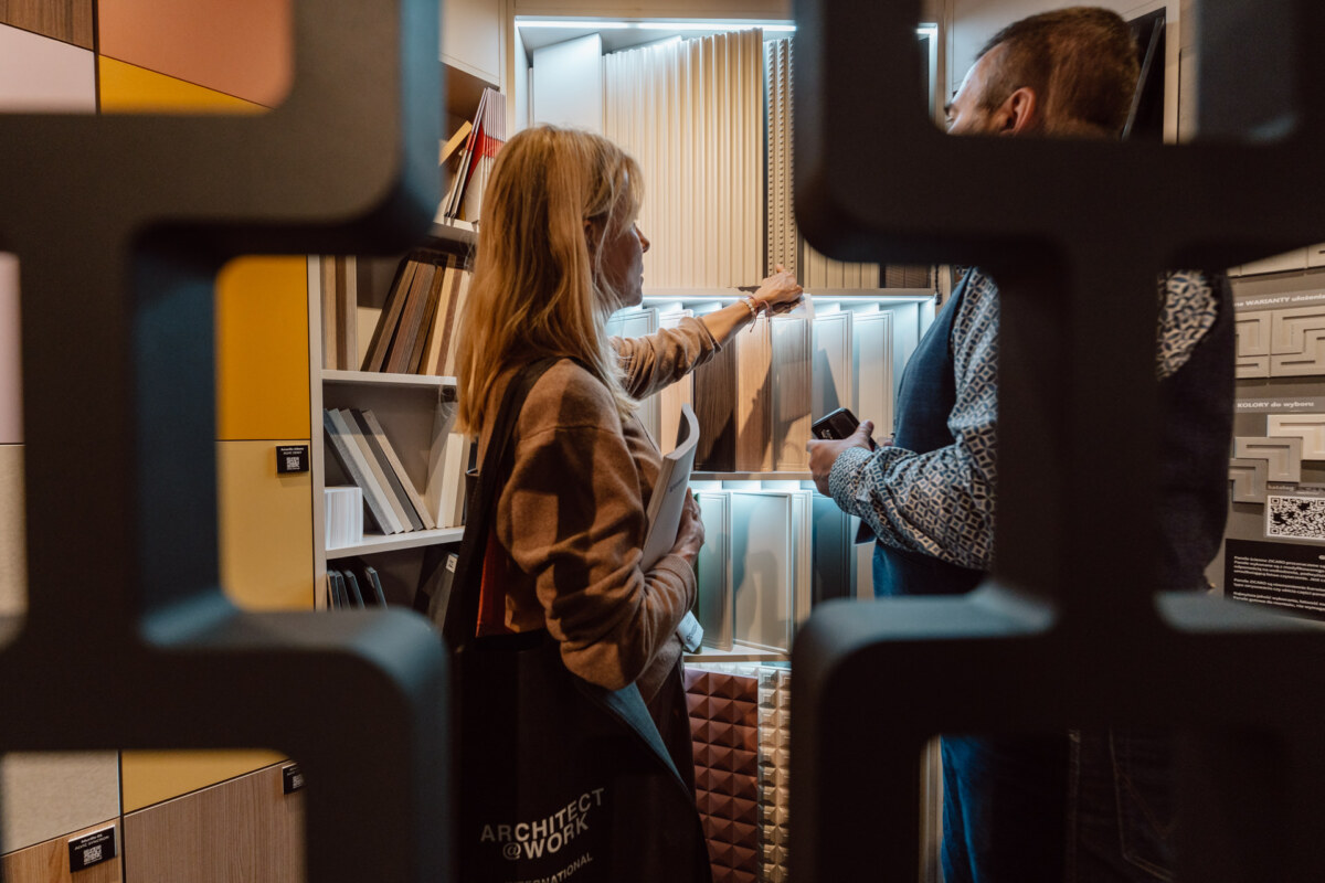 reportaz targi architektura architectatwork 2025 2 Zwei Personen stehen vor beleuchteten Wandpaneelen, diskutieren und begutachten Muster, die durch eine dekorative Trennwand zu sehen sind. Im Hintergrund sind Regale mit Mustern und verschiedenen Texturen zu sehen.