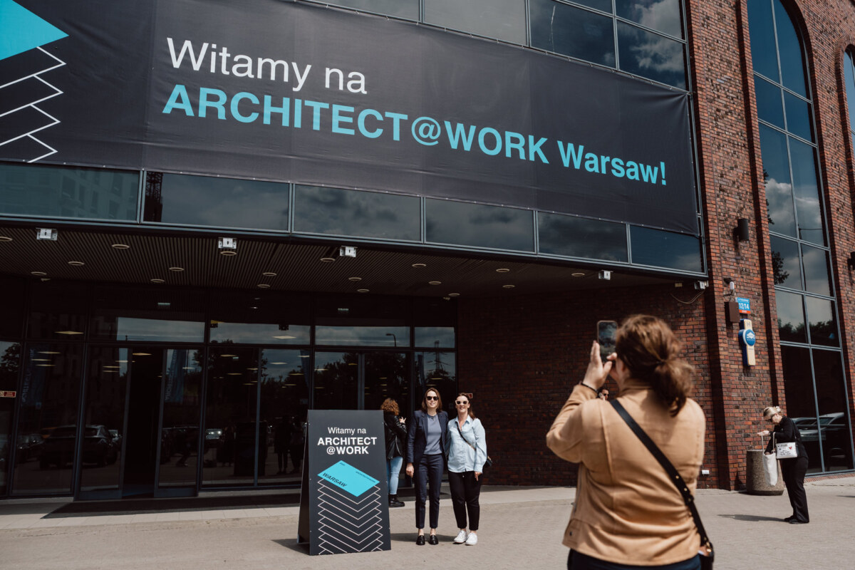 reportaz targi architektura architectatwork 2025 20 Eine Person macht ein Foto von drei Personen, die vor einem Gebäude mit einem großen Schild mit der Aufschrift "Welcome to ARCHITECT@WORK Warsaw!