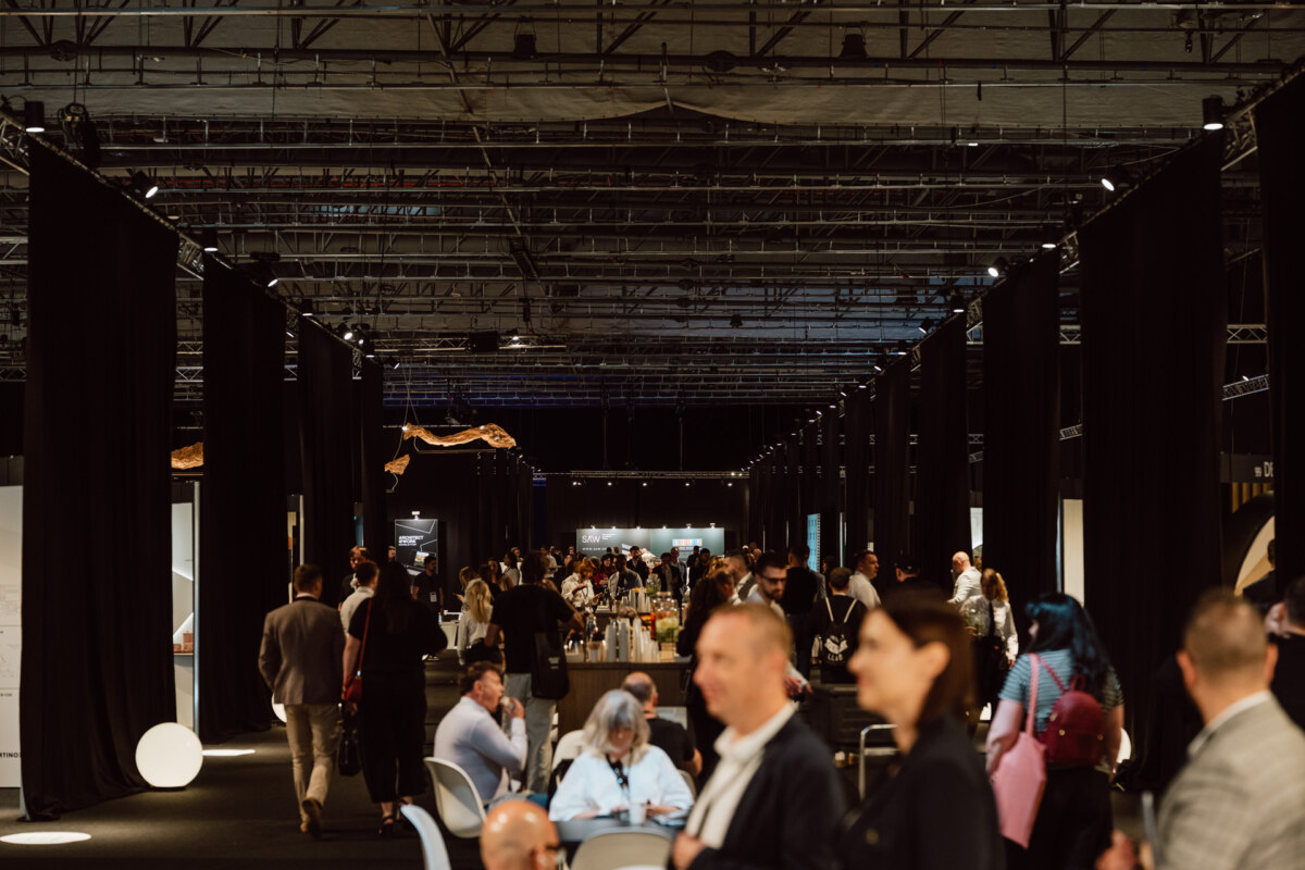 reportaz targi architektura architectatwork 2025 24 Eine große Gruppe von Menschen nimmt an der Veranstaltung in dem Gebäude mit den Ständen und Ausstellungen teil. Sie laufen und unterhalten sich in einer geräumigen Halle mit hohen schwarzen Vorhängen und einer industriellen Decke.