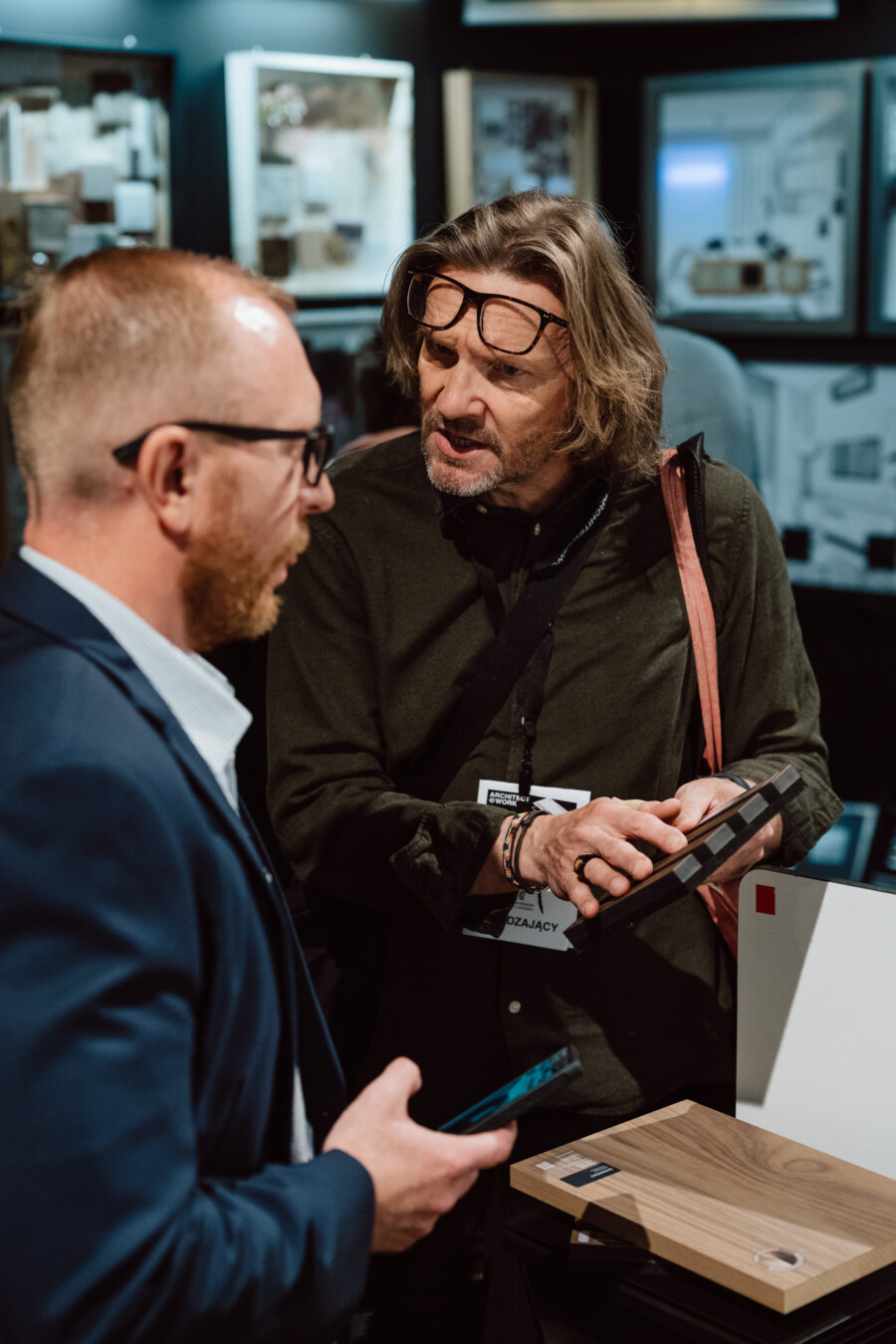 reportaz targi architektura architectatwork 2025 25 Zwei Männer unterhalten sich auf einer Veranstaltung oder Ausstellung; einer trägt eine Brille und hält ein Tablet, der andere trägt einen Anzug. Im Hintergrund sind Displays und Designmuster zu sehen.
