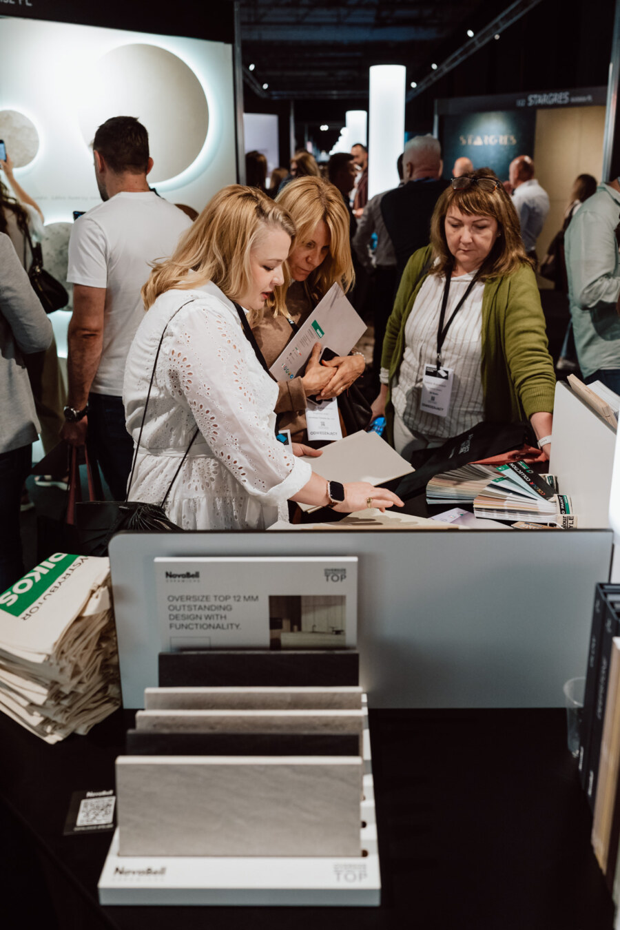 reportaz targi architektura architectatwork 2025 26 Drei Frauen untersuchen Materialproben an einem überfüllten Ausstellungsstand und diskutieren die verfügbaren Optionen. Andere Teilnehmer sind im Hintergrund zwischen den Auslagen und Schildern zu sehen. Im Vordergrund befindet sich ein Display mit Broschüren und Mustertafeln.
