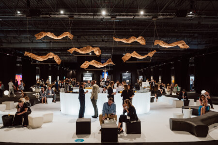 A modern room party with people gathering around a circular, lighted bar. Unique wooden sculptures hang from the ceiling, and attendees sit or stand in a spacious, stylish environment with black curtains and modern furniture.