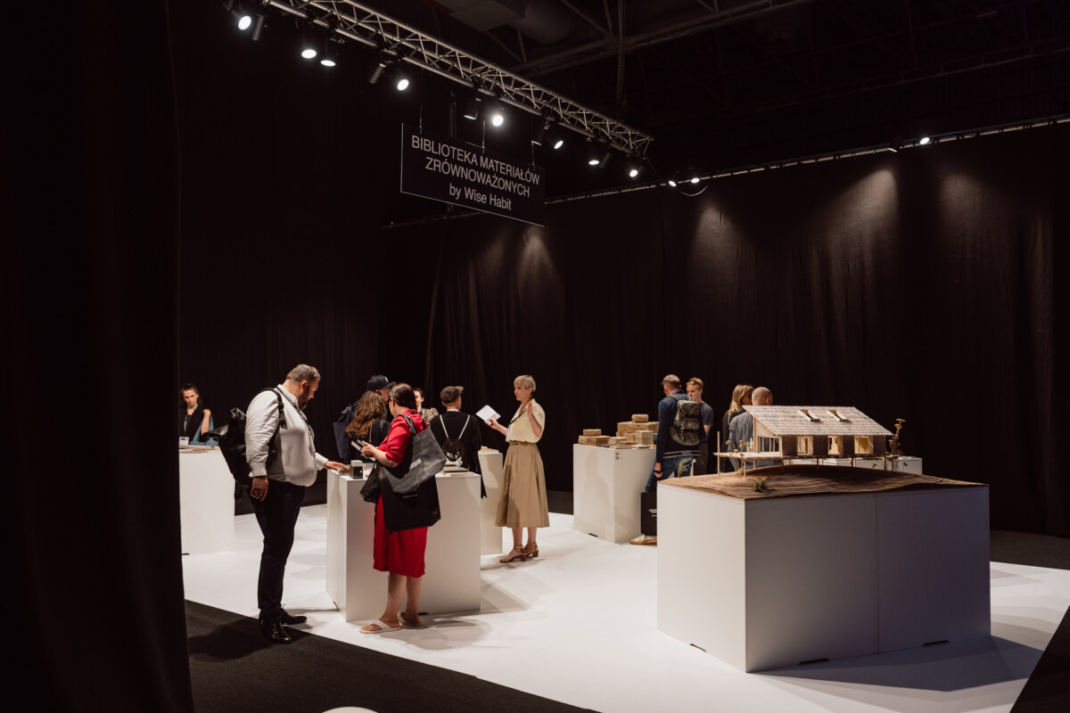 reportaz targi architektura architectatwork 2025 29 In einem schwach beleuchteten Museum oder einer Galerie mit schwarzen Wänden betrachten die Menschen die Exponate und interagieren. Auf den Ausstellungstischen stehen Modelle und Bücher. Oben steht auf einem Schild 'Wise Habit Sustainable Materials Library'.