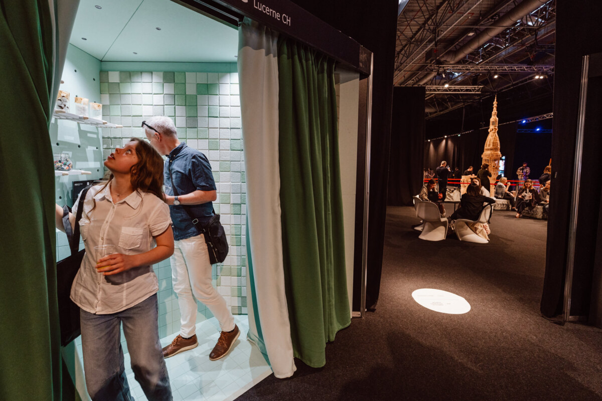 reportaz targi architektura architectatwork 2025 3 Eine Frau und ein Mann stehen in einem kleinen, gekachelten Messestand mit grünen Vorhängen. Die Frau schaut auf und hält ein Getränk in der Hand, der Mann steht hinter ihr. Im Hintergrund sitzen Menschen an Tischen in einem schwach beleuchteten Veranstaltungsraum.